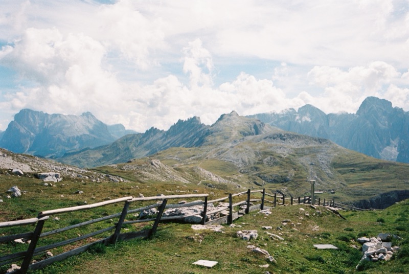 Frame 18: the Schlern above the Seiser Alm on a day of heavy cumulus. A weathered wooden fence runs down across the alpine grass in the foreground, and jagged Dolomite peaks stand along the horizon behind it.